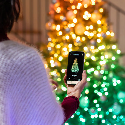 Person taking a photo of a Christmas tree with a smartphone in front of a blurred festive background